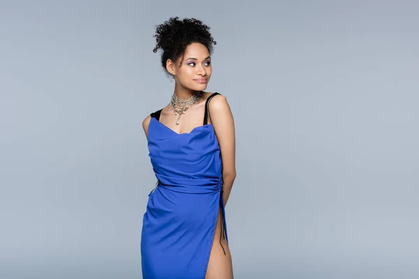 Femme afro-américaine souriante en robe bleu vif regardant loin tout en posant isolé sur gris — Photo de stock