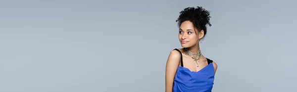 Smiling african american woman in bright blue dress looking away while posing isolated on grey, banner — Stock Photo