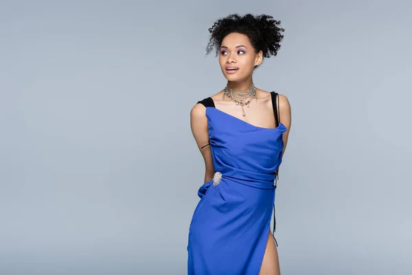 Curiosa mujer afroamericana en vestido azul brillante mirando hacia otro lado mientras posando aislada en gris - foto de stock