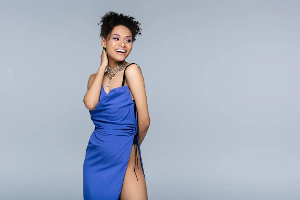 Heureuse femme afro-américaine en robe bleu vif regardant loin tout en posant isolé sur gris — Photo de stock