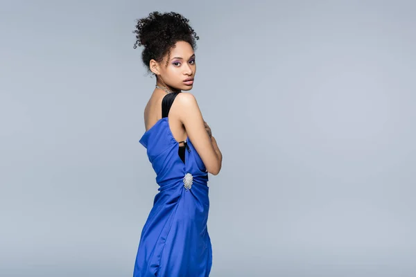 Encaracolado mulher americana africana em vestido de deslizamento azul olhando para a câmera enquanto posando isolado no cinza — Fotografia de Stock