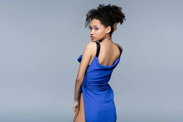 Joven africano americano mujer en brillante azul vestido mirando cámara mientras posando aislado en gris - foto de stock