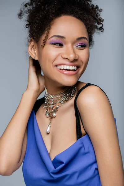 Счастливая африканская американка с пурпурной тенью для глаз, улыбающаяся изолированная на сером — стоковое фото