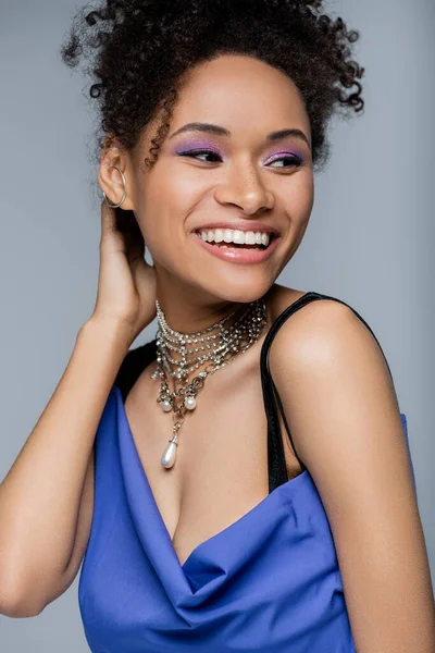 Happy african american woman with purple eye shadow smiling isolated on grey — Stock Photo