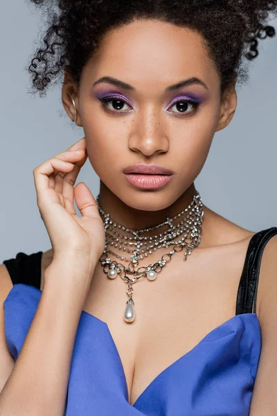 Curly african american woman with purple eye shadow touching face isolated on grey — Stock Photo