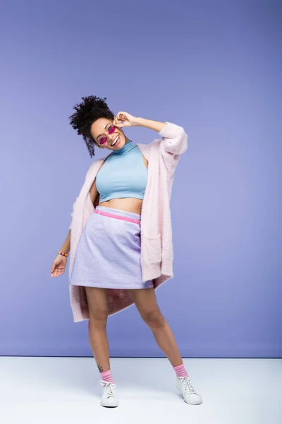 Full length of positive african american woman adjusting trendy sunglasses on violet — Stock Photo