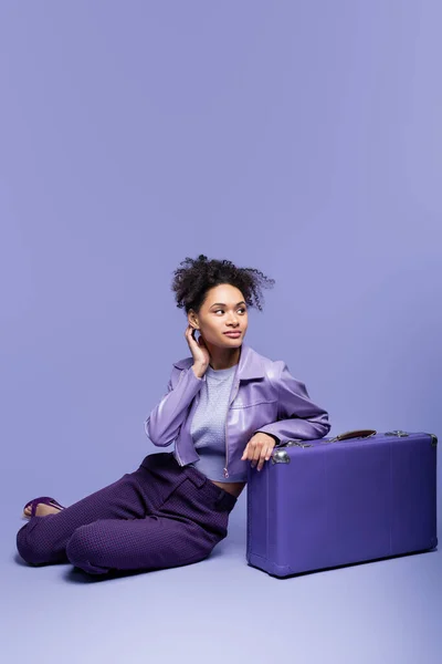 Повна довжина мрійливої афроамериканської моделі в шкіряній куртці, що сидить біля фіолетової валізи на фіолетовому — стокове фото