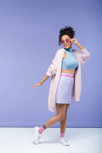 Full length of cheerful african american woman with tattoo adjusting trendy sunglasses on purple — Stock Photo