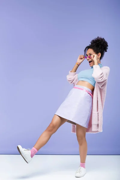 Longitud completa de mujer afroamericana asombrada ajustando gafas de sol rosadas en púrpura - foto de stock