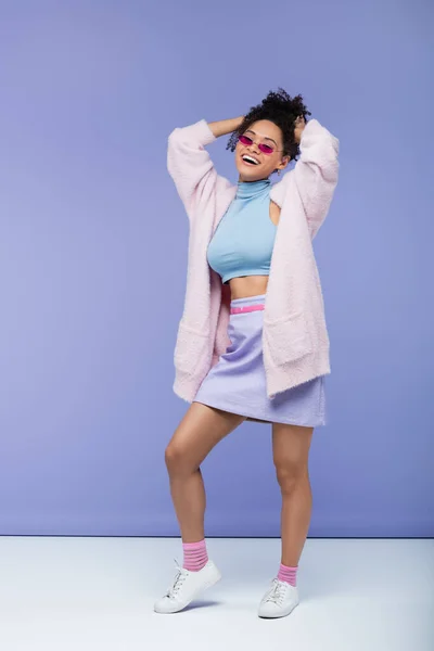 Longitud completa de la mujer americana africana alegre en gafas de sol elegantes que ajustan el pelo rizado en violeta - foto de stock