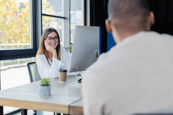 Cheerful businesswoman in glasses looking at blurred african american colleague in office — Stock Photo