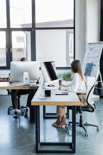 Joven mujer de negocios mirando a un colega afroamericano cerca de monitores de computadoras en la oficina - foto de stock