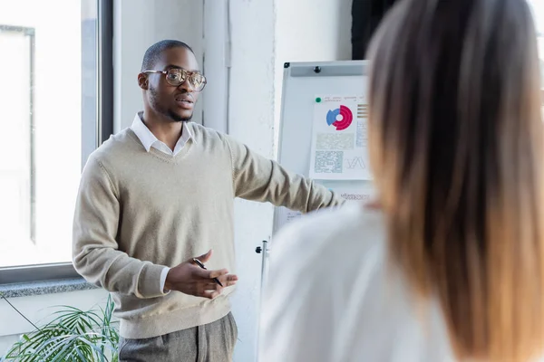 African american businessman in glasses talking to blurred colleague near flip chart — Stock Photo