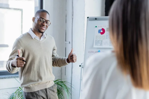 Heureux homme d'affaires afro-américain dans des lunettes montrant pouces vers le haut près de collègue flou et flip chart — Photo de stock