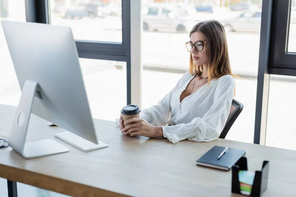 Femme d'affaires dans des lunettes tenant tasse en papier et regardant l'écran d'ordinateur dans le bureau — Photo de stock