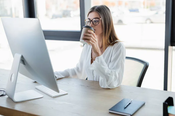 Young businesswoman in glasses drinking coffee to go and looking at computer monitor in office — Stock Photo
