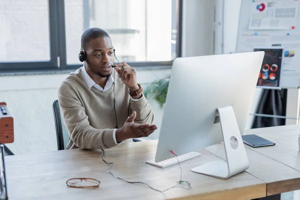 Opérateur afro-américain tenant le microphone tout en parlant et pointant vers le moniteur d'ordinateur — Photo de stock