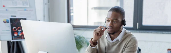 Operador afroamericano sosteniendo micrófono mientras habla y mirando el monitor de la computadora, pancarta - foto de stock