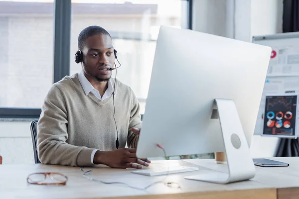 Operador afroamericano en auriculares hablando y mirando monitor de computadora - foto de stock