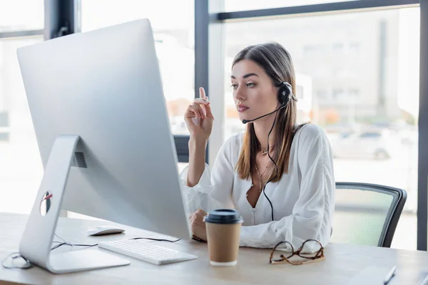 Empresária em fone de ouvido olhando para monitor de computador ao apontar com o dedo — Fotografia de Stock