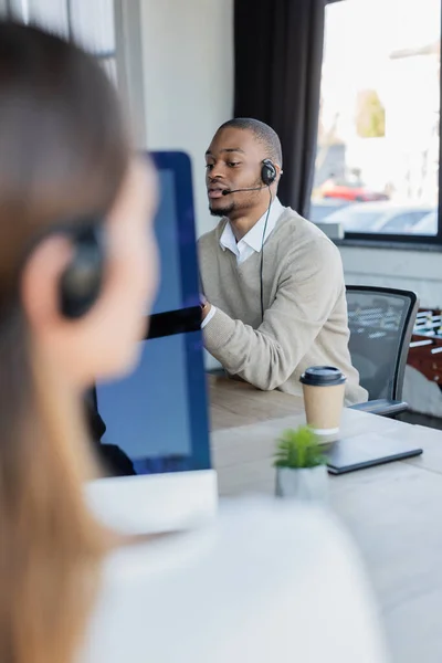 Africano americano operador en auriculares hablando cerca borrosa colega en la oficina - foto de stock