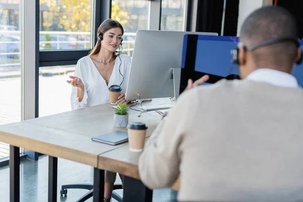 Happy businesswoman in headset looking at computer monitor near african american colleague — Stock Photo