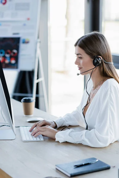 Side view of smiling businesswoman in headset typing on computer keyboard in office — Stock Photo