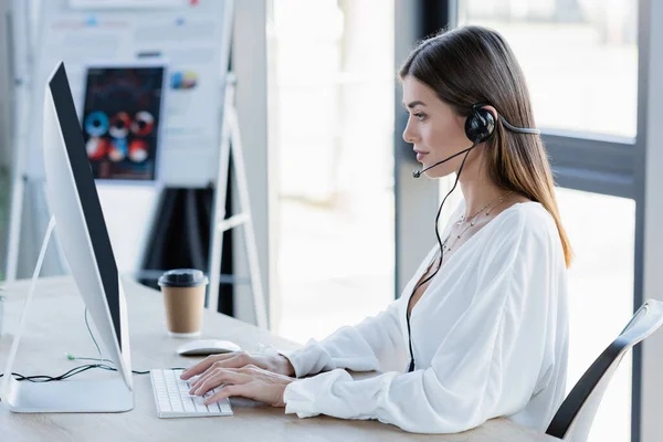 Side view of young woman in headset typing on computer keyboard in office — Stock Photo