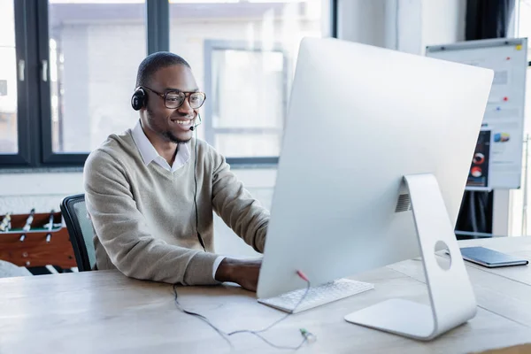 Glücklicher afrikanisch-amerikanischer Bediener mit Headset und Brille, der im Büro auf den Computermonitor blickt — Stockfoto