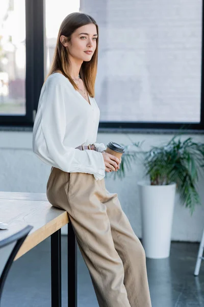 Pretty young businesswoman holding paper cup and leaning on desk in office — Stock Photo