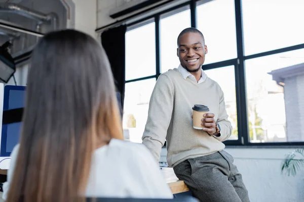Homme d'affaires afro-américain souriant tenant une tasse en papier et parlant avec un collègue flou — Photo de stock