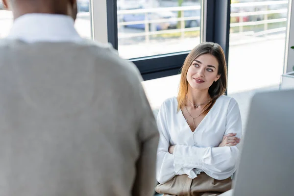 Fröhliche Geschäftsfrau sitzt in verschränkten Armen und blickt verschwommenen afrikanisch-amerikanischen Kollegen an — Stockfoto