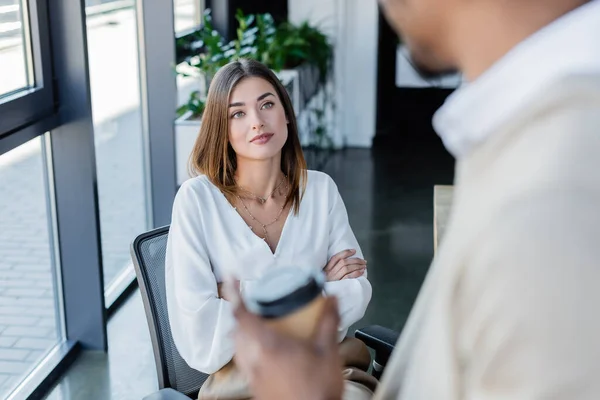 Young businesswoman sitting in crossed arms and looking at blurred african american colleague with paper cup — Stock Photo