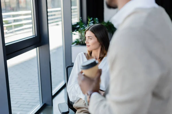 Femme d'affaires regardant fenêtre près floue confrère afro-américain avec tasse en papier — Photo de stock