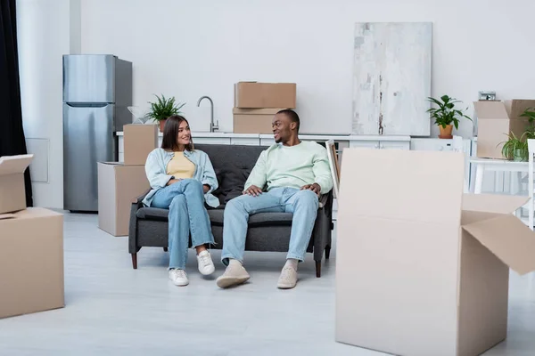 Happy interracial couple sitting on sofa around carton boxes during relocation — Stock Photo