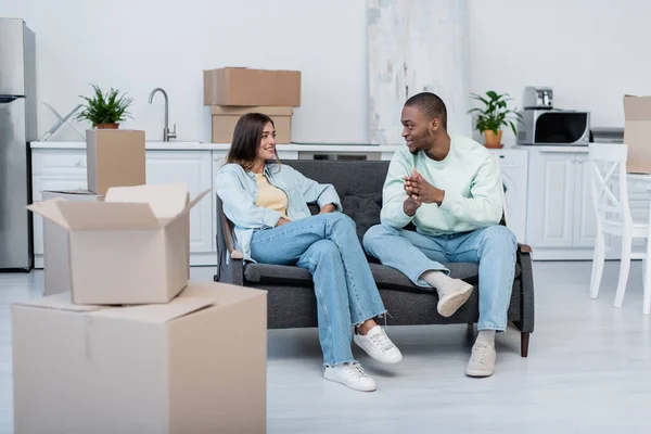 Cheerful interracial couple looking at each other while sitting on sofa near carton boxes during relocation — Stock Photo