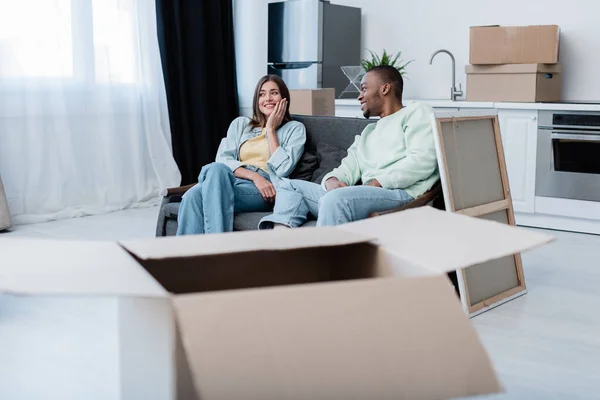 Fröhliches gemischtrassiges Paar sitzt während des Umzugs auf dem Sofa um Kartons — Stockfoto