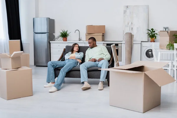 Tired interracial couple sitting on couch around carton boxes during relocation — Stock Photo