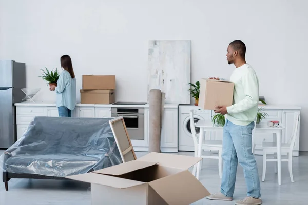 African american man holding carton box while looking at girlfriend with plant during relocation — Stock Photo