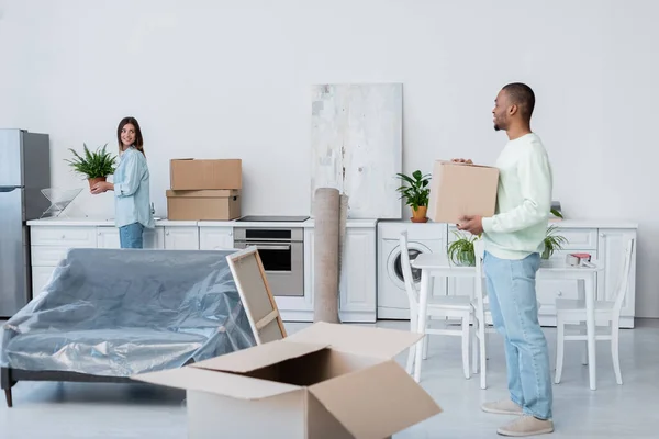 Happy african american man holding carton box while looking at girlfriend with plant during relocation — Stock Photo