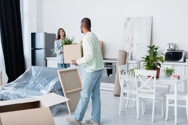 African american man holding carton box while looking at happy girlfriend with plant during relocation — Stock Photo
