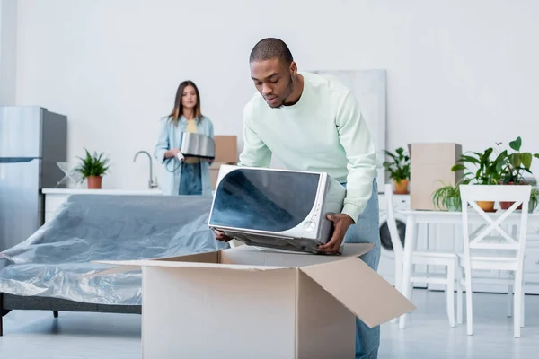 Homme afro-américain mettant four à micro-ondes dans la boîte en carton pendant l'emballage lors de la relocalisation — Photo de stock