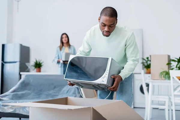 African american man putting microwave oven in carton box during relocation — Stock Photo