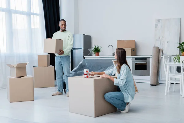 Young woman holding adjusting tape and packing carton box near african american man in living room — Stock Photo