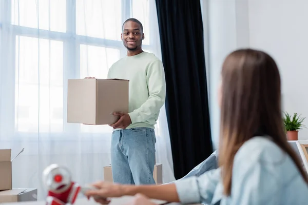 Africano americano hombre holding caja y mirando borrosa mujer en casa - foto de stock