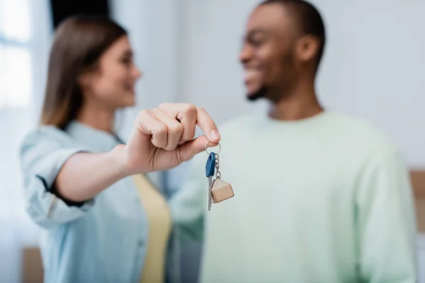 Mujer borrosa y feliz sosteniendo la llave cerca de hombre afroamericano en nuevo apartamento - foto de stock