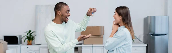 Hombre afroamericano feliz sosteniendo la llave cerca de novia excitada en nuevo apartamento, pancarta - foto de stock