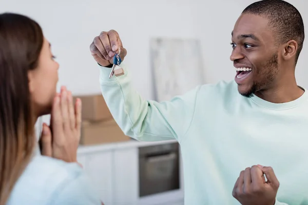 Hombre afroamericano feliz sosteniendo la llave cerca de novia borrosa en nuevo apartamento - foto de stock