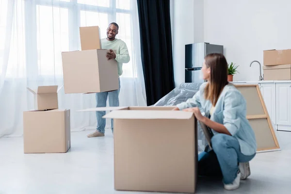 Happy african american man holding boxes near girlfriend in new apartment — Stock Photo