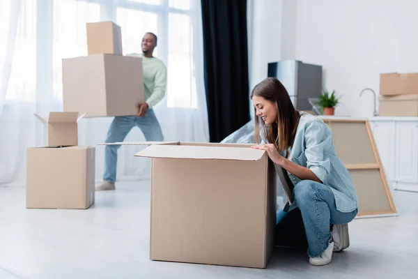 Feliz jovem mulher desempacotando caixa perto de homem americano africano borrado em novo apartamento — Fotografia de Stock